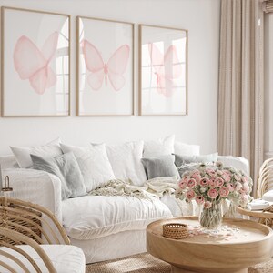 May include: Three framed prints of pink watercolor butterflies on a white wall above a white couch with gray pillows and a wooden coffee table with a vase of pink flowers.