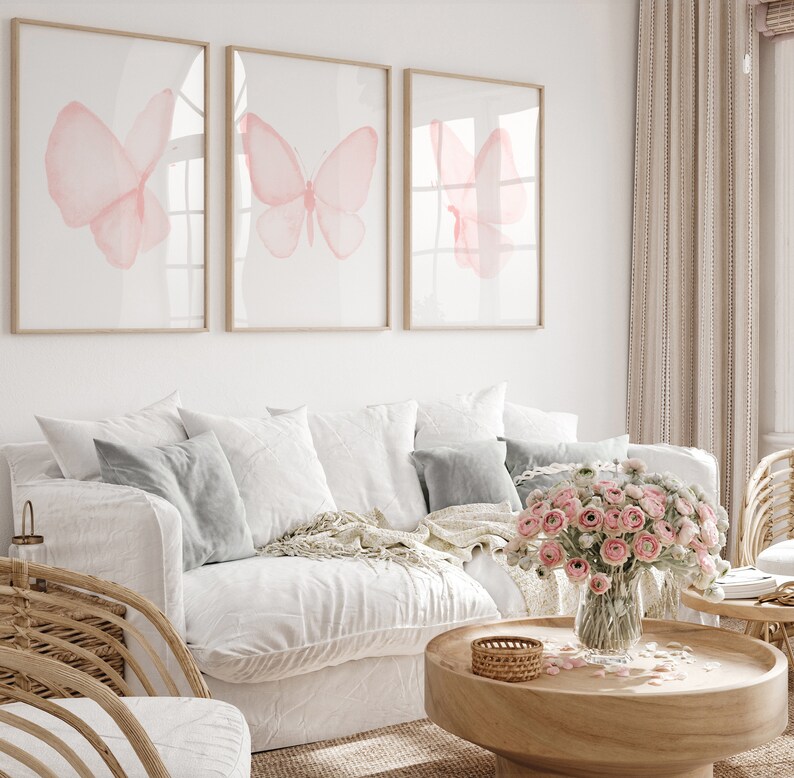 May include: Three framed prints of pink watercolor butterflies on a white wall above a white couch with gray pillows and a wooden coffee table with a vase of pink flowers.
