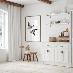 May include: A white kitchen with a white cabinet, a white countertop, and a gold framed print of two black birds in flight. The print is hanging on a white wall. There are two small wooden stools in front of the cabinet.