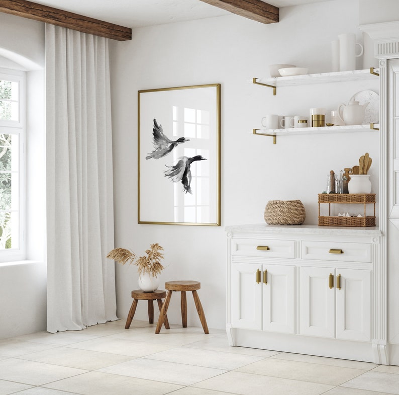 May include: A white kitchen with a white cabinet, a white countertop, and a gold framed print of two black birds in flight. The print is hanging on a white wall. There are two small wooden stools in front of the cabinet.