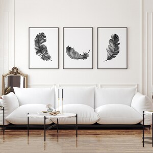 K&ouml;nnte beinhalten: Drei Schwarz-Wei&szlig;-Aquarell-Federdrucke in schwarzen Rahmen an einer wei&szlig;en Wand &uuml;ber einem wei&szlig;en Sofa.