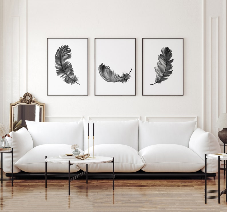 K&ouml;nnte beinhalten: Drei Schwarz-Wei&szlig;-Aquarell-Federdrucke in schwarzen Rahmen an einer wei&szlig;en Wand &uuml;ber einem wei&szlig;en Sofa.