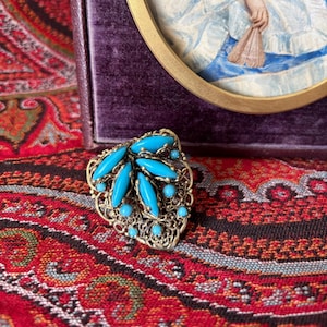 May include: A turquoise and gold filigree brooch with a leaf design. The brooch is on a red and black paisley patterned fabric.