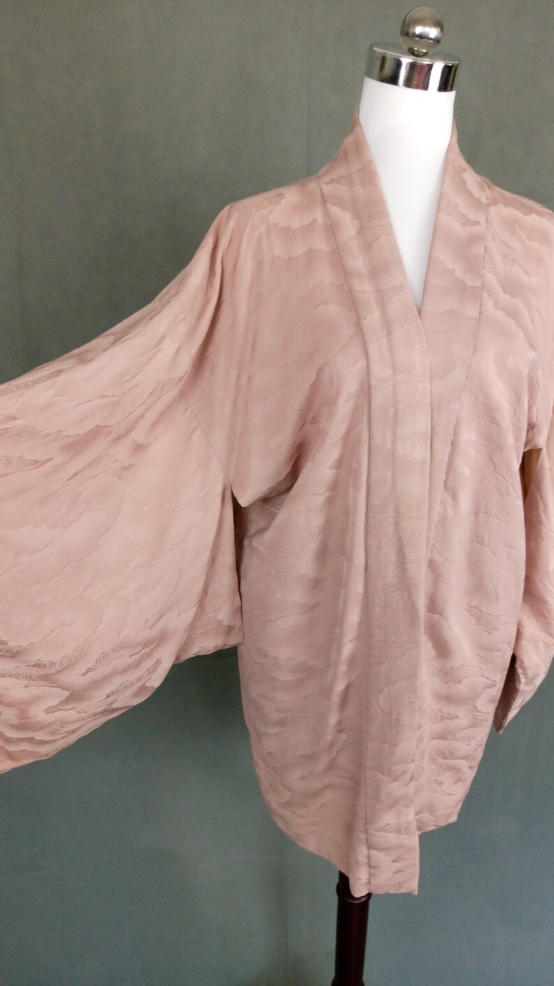 May include: A pale pink kimono with a subtle cloud pattern. The kimono is made of a soft, silky fabric and has long, wide sleeves.