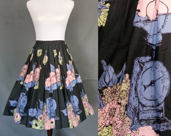 May include: A black full skirt with a floral print featuring pink, blue, and green flowers. The skirt has a pleated design and a waistband. The print also includes a vintage scale with the text "Candy Scale Chicago".