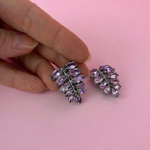 May include: A pair of purple crystal leaf earrings. The earrings are made of silver metal and have a delicate, feminine design.