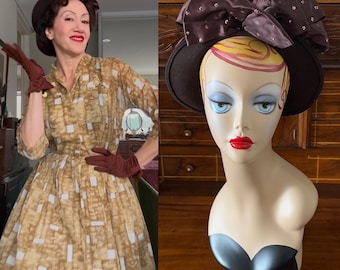 Puede incluir: Un sombrero marrón con un lazo grande, exhibido en una cabeza de maniquí. El lazo tiene un estampado de lunares. El maniquí tiene ojos azules y lápiz labial rojo. El sombrero es de estilo vintage, adecuado para ocasiones especiales.