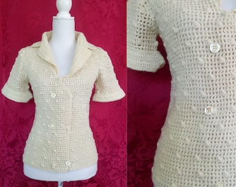 May include: A cream-colored crocheted top with a collar and short sleeves. The top has a button-up front and a textured design.