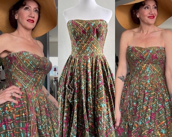May include: A strapless, olive green vintage dress with a colorful grid pattern of pink, orange, and blue lines. The dress has a full skirt and a fitted bodice. A wide-brimmed straw hat completes the look.