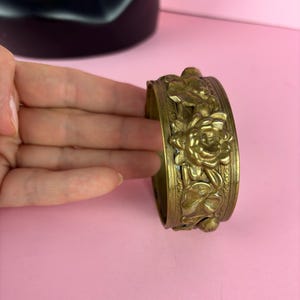 May include: A gold-toned metal bracelet with a floral design. The bracelet is made of a thick metal and has a detailed design of flowers and leaves.