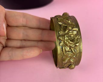 Puede incluir: Un brazalete de metal dorado con un diseño floral. El brazalete está hecho de un metal grueso y tiene un diseño detallado de flores y hojas.