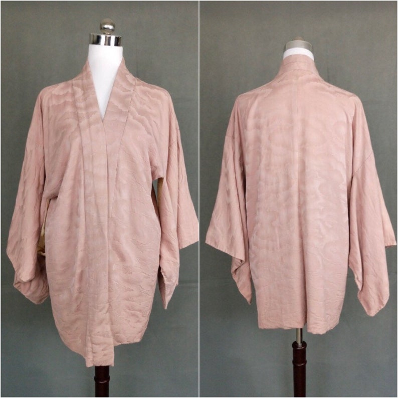 May include: A pale pink Japanese kimono with a subtle wave pattern. The kimono is made of a soft, silky fabric and has long sleeves.