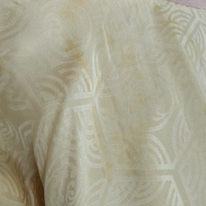 May include: Close-up of a cream-colored fabric with a subtle embossed pattern. The fabric has a few light brown stains.