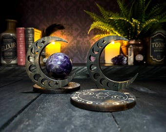 May include: Two wooden crescent moon candle holders with a purple amethyst sphere in the center of each. The holders are on a dark wooden surface.