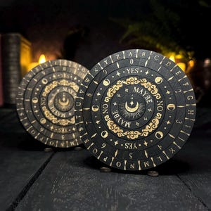May include: Two circular, wooden divination boards. One is black with gold lettering and a crescent moon design, the other is brown with a similar design. The boards have the words "YES", "MAYBE", and "NO" and are used for spiritual practices.