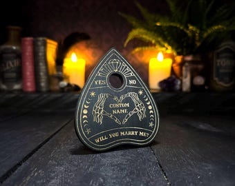 May include: A black wooden planchette featuring gold-colored engravings of a diamond, moon phases, and two skeleton hands forming a heart shape. The text reads YES, NO, CUSTOM NAME, and WILL YOU MARRY ME? with a circular hole at the top center.