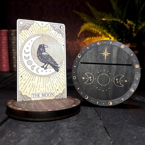 May include: A rectangular tarot card titled The Moon featuring a black raven perched on a crescent moon, displayed in a circular dark wood stand. Beside it sits a matching circular dark wood stand engraved with a star and moon phase symbols.
