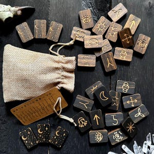 May include: A set of wooden divination runes with various symbols and designs, including moons, stars, and arrows. The runes are arranged on a dark wooden surface and come in a small hessian bag.