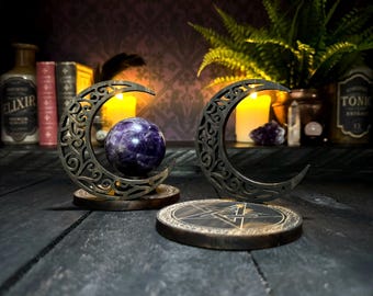 May include: Two dark wooden crescent moon stands with intricate scrollwork cutouts. One stand holds a polished purple amethyst sphere. The other stand rests on a circular wooden base featuring an engraved pentagram design with fine line details.