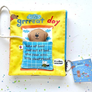 May include: A yellow fabric book with a brown dog peeking out of a bed. The book has the text "Gus's grrreat day" on the cover and the text "Wake up Gus! Jump out of bed. open your eyes. little sleepyhead." on a blue fabric flap. The book has a small tag attached to it that says "GUS'S grrreat day".