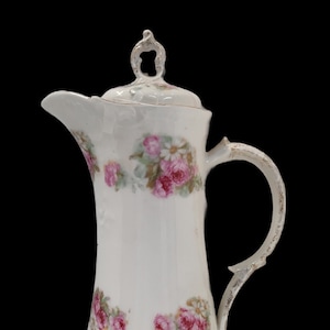 May include: White porcelain pitcher with a floral design featuring pink roses and green leaves. The pitcher has a lid with a decorative knob and a handle with gold accents.