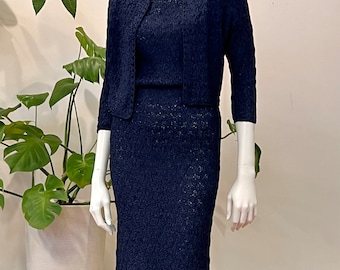 May include: A navy blue knit dress and matching cardigan set. The dress has a fitted silhouette and a slightly flared hem. The cardigan has a cropped length and a round neckline.