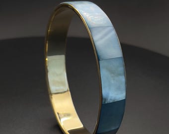 Puede incluir: Una pulsera rígida circular con un interior en tono dorado pulido y un exterior liso incrustado con segmentos rectangulares de nácar azul iridiscente. La pulsera tiene un diseño de banda sólida y continua con un acabado reflectante.