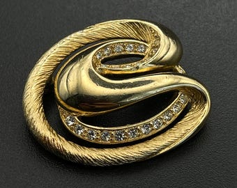 Puede incluir: Un broche de tono dorado con un diseño ovalado abstracto y en espiral. La pieza combina un acabado de metal cepillado texturizado con secciones doradas lisas y pulidas, y una fila curva de pequeños diamantes de imitación claros y brillantes.