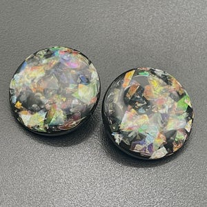 Könnte beinhalten: Zwei runde, schwarze Ohrringe mit einem Mosaik aus bunten, irisierenden Teilen, die in die Oberfläche eingebettet sind. Die Ohrringe haben eine glänzende Oberfläche und einen dunklen Hintergrund.