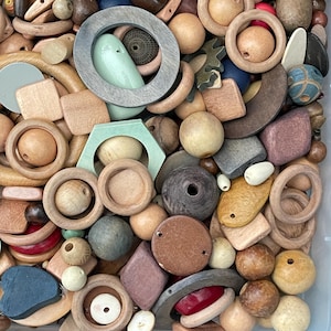 May include: A collection of assorted wooden beads and shapes in various sizes and colors, including natural wood, grey, green, blue, red, and brown tones. The assortment features rings, spheres, squares, and geometric pieces with drilled holes.