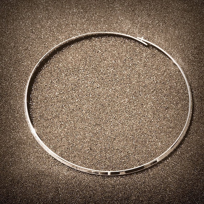 May include: A silver-colored, circular necklace with a clasp. The necklace is a thin, smooth band, and it is set against a textured, dark background.