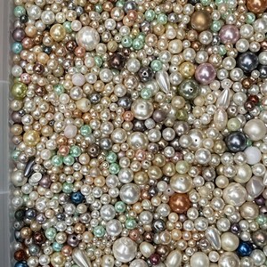 May include: A large collection of loose beads in various sizes and shapes, featuring a mix of white, cream, gold, silver, bronze, pink, and light green colors. The beads have smooth, lustrous finishes, with some spherical and some teardrop shapes.