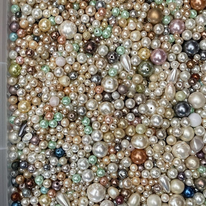 May include: A large collection of loose beads in various sizes and shapes, featuring a mix of white, cream, gold, silver, bronze, pink, and light green colors. The beads have smooth, lustrous finishes, with some spherical and some teardrop shapes.