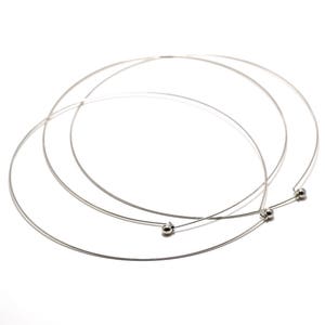 May include: Three silver-colored choker necklaces with a minimalist design. Each necklace is a thin, circular wire with a small, round bead at the clasp. The necklaces are displayed on a white background.