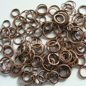 Op de afbeelding: Een verzameling van talrijke koperkleurige metalen buigringen in verschillende maten. Elke ring is cirkelvormig met een kleine opening en verspreid over het kader. Het metaal heeft een warme, bruinachtige metallic afwerking.