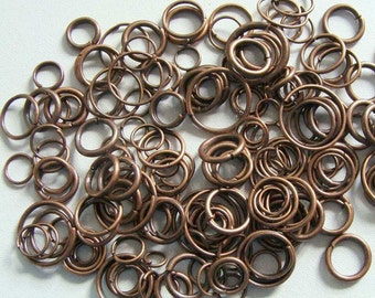 May include: A collection of numerous copper-toned metal jump rings in various sizes. Each ring is circular with a small split opening, scattered across the frame. The metal has a warm, brownish-metallic finish with a smooth, rounded wire texture.