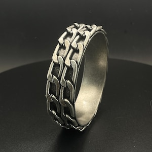 Könnte beinhalten: Ein silberfarbener Ring mit einem Kettenmuster. Der Ring hat drei Reihen ineinandergreifender Kettenglieder um das Band. Der Ring besteht aus Metall mit mattem Finish und ist vor dunklem Hintergrund platziert.
