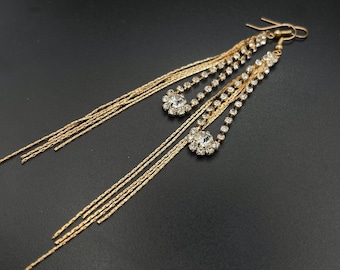 Puede incluir: Un par de pendientes largos colgantes en tono dorado con múltiples cadenas delicadas y dos detalles circulares incrustados con brillantes transparentes. Los pendientes cuentan con un cierre de gancho para su colocación.