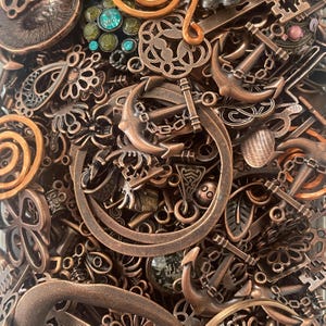 Pode incluir: Uma coleção de amuletos e enfeites de metal de cor bronze antigo. A variedade inclui âncoras, chaves, filigranas e elementos circulares decorativos. Alguns amuletos têm detalhes de joias turquesas e verdes.