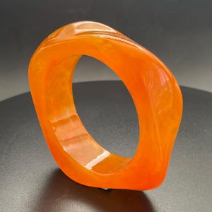 Könnte beinhalten: Ein orangefarbenes, durchscheinendes Armreif-Armband. Das Armband hat eine abgerundete, quadratische Form mit glatten Kanten. Die Farbe ist ein leuchtendes Orange mit einigen helleren, marmorierten Bereichen. Das Armband besteht aus einem festen Material und scheint ein Statement-Stück zu sein.