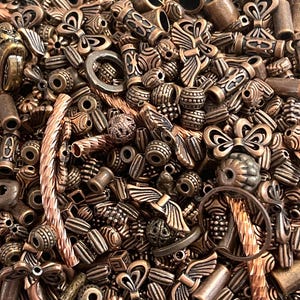 Könnte beinhalten: Eine große Auswahl an kupferfarbenen Metallperlen in verschiedenen Formen und Größen. Die Perlen umfassen Flügel, Schmetterlinge und runde Perlen mit komplizierten Designs.