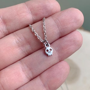 May include: A silver-colored necklace featuring a small skull pendant. The skull has two eye sockets and a nose cavity. The chain is delicate and appears to be made of small, linked metal pieces. The necklace is held in a hand.