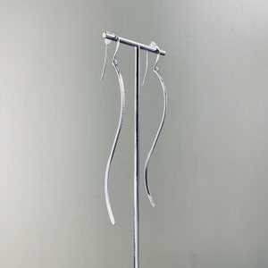 May include: A pair of silver earrings with a curved, wavy design. The earrings are hanging from a stand.