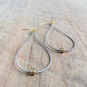 May include: A pair of teardrop-shaped earrings made from silver wire with gold accents. The earrings are suspended from a gold hook.