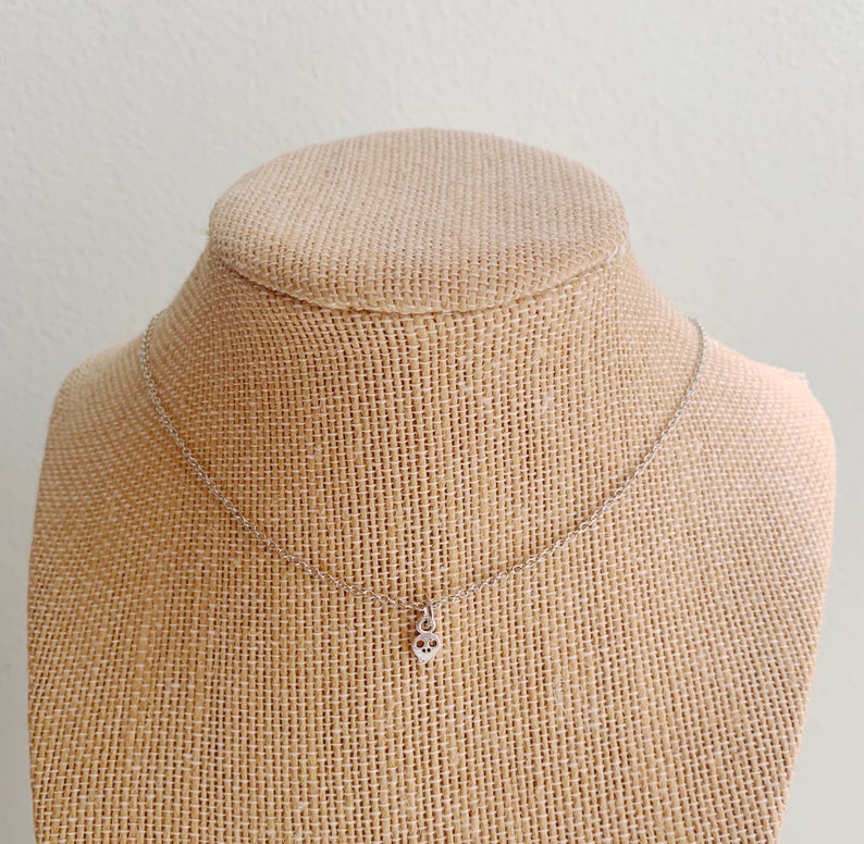 May include: A silver-colored necklace with a small skull pendant. The pendant is detailed with eye sockets and a nose cavity. The necklace is displayed on a burlap-covered bust.
