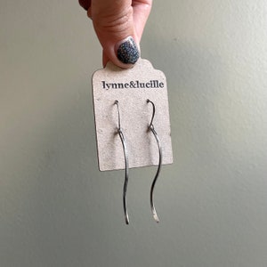 May include: A pair of silver-toned, wavy drop earrings. The earrings are displayed on a brown tag with the text "lynne&lucille" printed in black. The earrings have a textured design on one side.