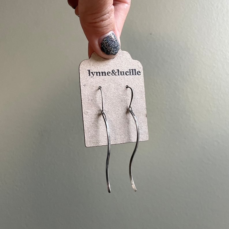 May include: A pair of silver-toned, wavy drop earrings. The earrings are displayed on a brown tag with the text "lynne&lucille" printed in black. The earrings have a textured design on one side.