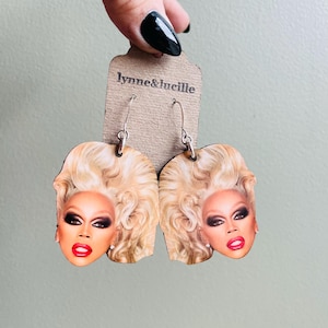 May include: A pair of wooden earrings featuring a printed image of a blonde woman with red lipstick. The earrings are on a brown card with the text "lynne&lucille" printed on it.
