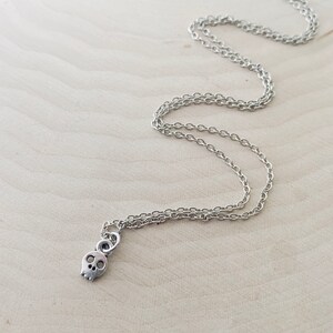 May include: Silver-toned necklace featuring a small skull pendant. The skull has two eye sockets and a nose cavity. The chain is a delicate link style. The necklace is displayed on a light-colored wooden surface.