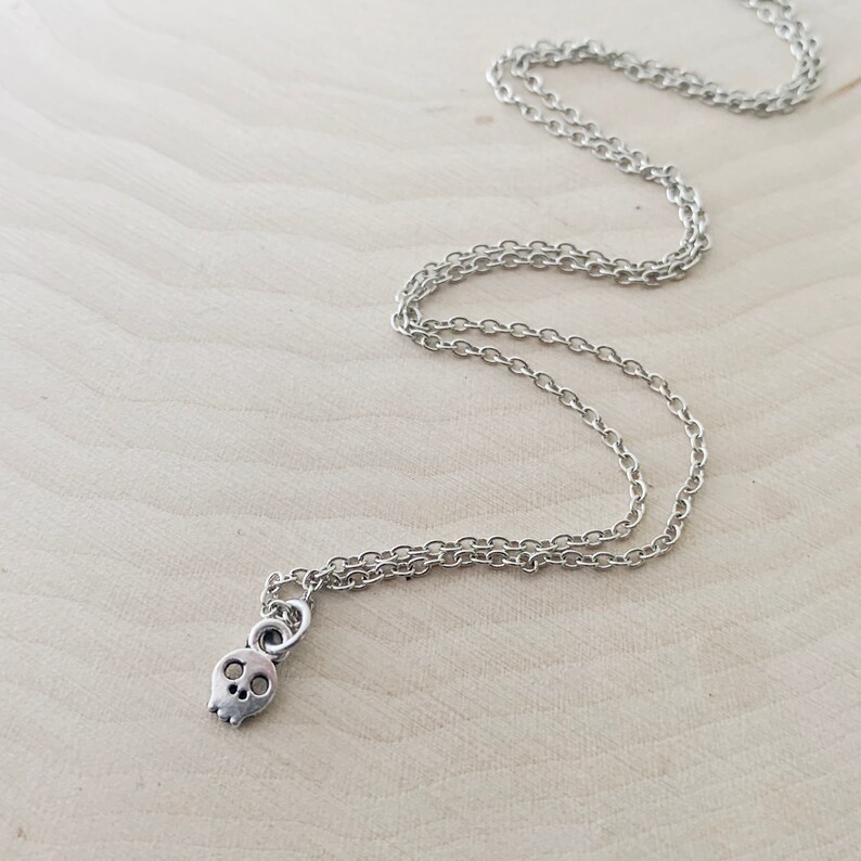 May include: Silver-toned necklace featuring a small skull pendant. The skull has two eye sockets and a nose cavity. The chain is a delicate link style. The necklace is displayed on a light-colored wooden surface.